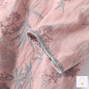 May include: Close-up of a pink fabric with a floral pattern in gray and purple. The sleeve of a garment is visible, with a gray trim along the edge. The fabric has a textured appearance.