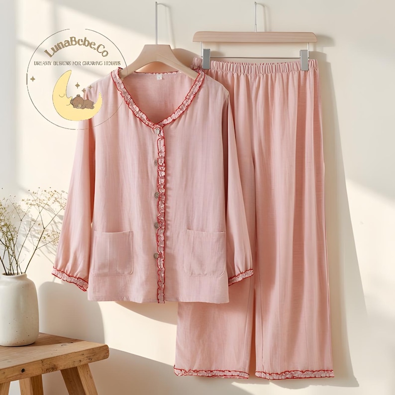 May include: A light pink pajama set with a long-sleeved button-down top and matching pants. The top has a ruffled trim in red along the collar, button line, and cuffs. The pants have a matching red trim at the hem. The set is hanging on a wooden hanger.