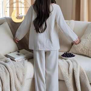 May include: A white pajama set with red trim, consisting of a long-sleeved top and pants. The top has a round neckline and the cuffs and hem are trimmed with red. The pajama set is made of a soft, lightweight fabric.
