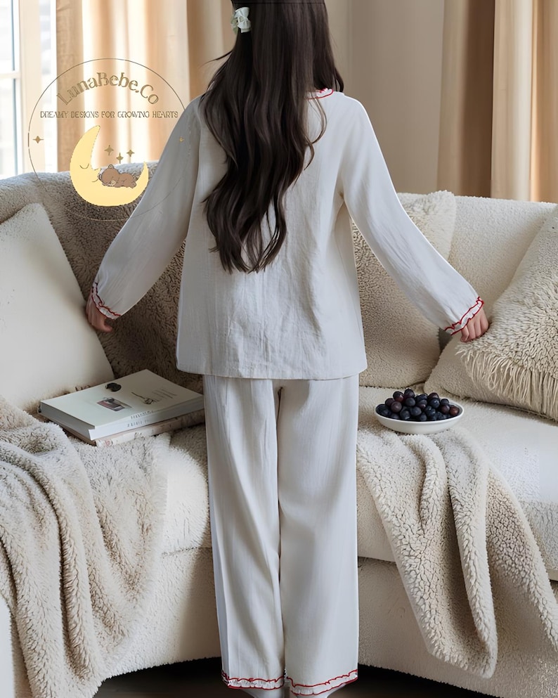 May include: A white pajama set with red trim, consisting of a long-sleeved top and pants. The top has a round neckline and the cuffs and hem are trimmed with red. The pajama set is made of a soft, lightweight fabric.