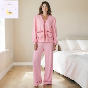 Puede incluir: Un conjunto de pijama rosa con una parte superior con cuello en V con bolsillos en forma de corazón y pantalones de pierna ancha a juego. La parte superior y los pantalones tienen ribetes rojos. El pijama está hecho de un material suave. El conjunto está diseñado para la comodidad y la relajación.