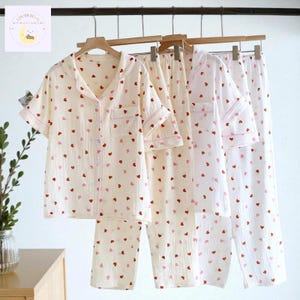Può includere: Set di pigiami color crema con motivi a cuore rossi e rosa appesi a grucce di legno. Ogni set include una camicia abbottonata a maniche corte e pantaloni abbinati. I pigiami hanno finiture rosa e sono realizzati in tessuto leggero.