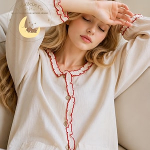 May include: Cream-colored pajama top with red ruffled trim around the collar, cuffs, and button placket. The top has a button-down front and a pocket. The Luna Bebe Co. logo is in the upper left corner.