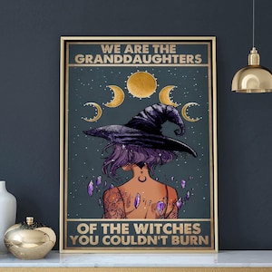 May include: A rectangular wall poster in a gold frame featuring a purple-haired figure wearing a black witch hat. Above the figure are gold moon phases and below are floating purple crystals. The text reads WE ARE THE GRANDDAUGHTERS OF THE WITCHES YOU COULDN'T BURN.