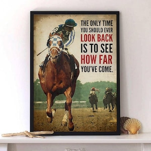 May include: A framed print depicting a horse race. A jockey in blue and white attire rides a brown horse in the lead. The print displays the text: "THE ONLY TIME YOU SHOULD EVER LOOK BACK IS TO SEE HOW FAR YOU'VE COME."