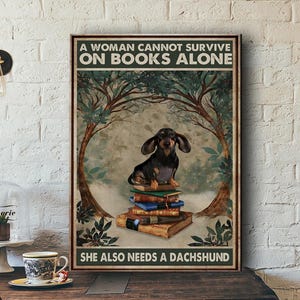 Peut inclure: Une affiche encadr&eacute;e avec un teckel assis sur une pile de livres. Le texte dit : &laquo; UNE FEMME NE PEUT PAS SURVIVRE SEULE AVEC DES LIVRES. ELLE A AUSSI BESOIN D'UN TECKEL &raquo;. L'&oelig;uvre a un style vintage avec une palette de couleurs marron et vert.