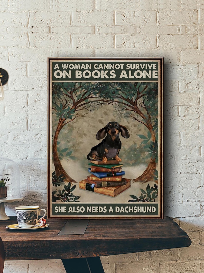 Peut inclure: Une affiche encadr&eacute;e avec un teckel assis sur une pile de livres. Le texte dit : &laquo; UNE FEMME NE PEUT PAS SURVIVRE SEULE AVEC DES LIVRES. ELLE A AUSSI BESOIN D'UN TECKEL &raquo;. L'&oelig;uvre a un style vintage avec une palette de couleurs marron et vert.