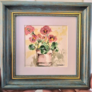 Peut inclure: Une aquarelle représentant des fleurs roses dans un pot rose clair, encadrée dans un cadre carré à la finition bleue vieillie avec une bordure intérieure dorée. L'œuvre présente un feuillage vert et un fond jaune, signée RRDANIELS dans le coin.