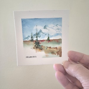 May include: A small watercolour painting of a mountain landscape. The artwork depicts snow-capped mountains under a blue sky with clouds. The foreground features trees and a field of brown and green. The artist's signature, "RR Daniels," is visible.