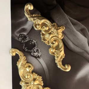 May include: Two gold-colored metal decorative embellishments with intricate floral designs. The embellishments are shaped like a stylized letter 'S' and have a raised, textured surface.