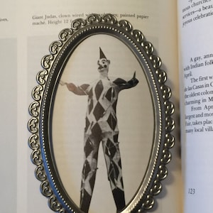 May include: A black and white photo of a clown in a diamond patterned costume, standing with arms outstretched. The photo is framed in a silver oval frame with a decorative border.