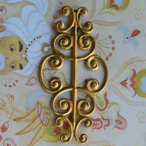 May include: A gold-colored, ornate metal decorative element with a symmetrical design. The piece features a central vertical bar with swirling scrollwork on either side. The background is a floral pattern in shades of yellow, cream, and red.