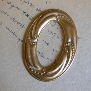 May include: Oval-shaped, antique gold-colored frame with ornate detailing. The frame features a decorative border with small, raised dots and swirling, stylized floral elements. The background is a light-colored paper with handwritten script.