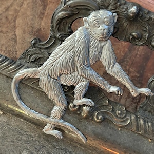 May include: A silver tray with an ornate monkey design. The monkey is sitting on a decorative scrollwork and has a long tail that wraps around the tray.