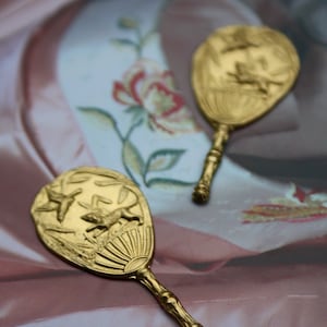 Puede incluir: Dos espejos de mano antiguos de color dorado. Cada espejo presenta un diseño en relieve detallado de pájaros, una rana y follaje. Los mangos tienen una textura similar al bambú. Los espejos están sobre un fondo con un patrón floral.