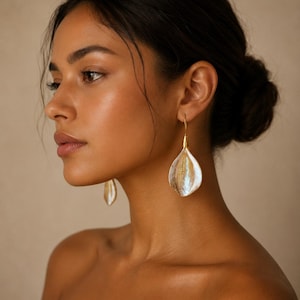 May include: A pair of drop earrings featuring a teardrop-shaped, iridescent white shell-like component suspended from a gold-toned metal hook. The earrings have a smooth, curved surface that reflects light, creating a pearlescent sheen.