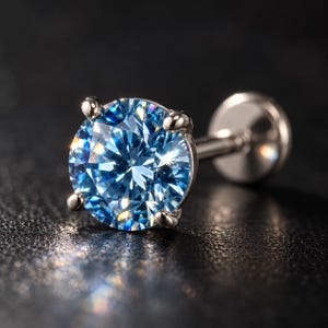 May include: A round, brilliant-cut light blue gemstone set in a four-prong silver-toned metal setting. The jewelry piece features a flat circular back plate attached to a straight post, designed as a stud earring or body piercing jewelry.