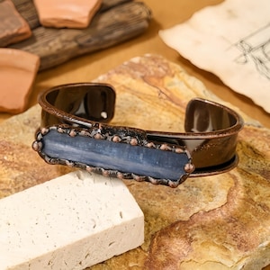 May include: A copper-toned cuff bracelet with a rectangular, blue-grey stone centerpiece. The bracelet has a dark, glossy finish and a textured border around the stone. The cuff is displayed on a natural stone surface.