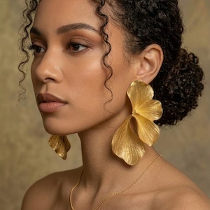 Könnte beinhalten: Goldfarbene Statement-Ohrringe in floralem Design. Die Ohrringe sind groß und strukturiert, mit einem geschichteten Blütenblatt-Aussehen. Eine zarte Goldkette ist ebenfalls sichtbar. Das Bild ist vor einem neutralen Hintergrund aufgenommen.