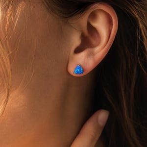 Könnte beinhalten: Ein dreieckiger Ohrstecker mit einem leuchtend blauen Opalstein, der irisierende Farbflecken aufweist. Der Stein ist in einer einfachen Metallfassung mit Krappen befestigt, was für ein klares, geometrisches Aussehen direkt am Ohrläppchen sorgt.