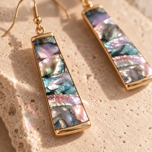 May include: A pair of gold-toned drop earrings featuring elongated rectangular pendants inlaid with iridescent abalone shell fragments. The shell pieces display a mosaic of shimmering purple, blue, and green hues, suspended from simple gold-toned hooks.