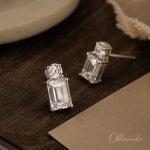 May include: A pair of silver-toned stud earrings, each featuring a clear, rectangular emerald-cut stone positioned below a smaller, round-cut clear stone. The earrings are secured with a post backing and display a bright, reflective, faceted surface.