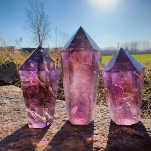 May include: Three amethyst crystal points with faceted tops are displayed outdoors. The crystals range in size and exhibit a gradient of purple and pink hues. They are set on a stone surface with a backdrop of grass, trees, and a bright blue sky.