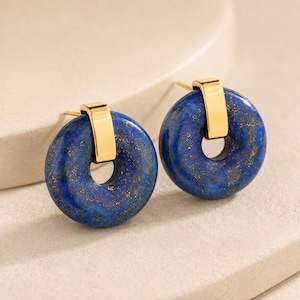 Könnte beinhalten: Ein Paar Ohrstecker mit kreisförmigen Lapislazuli-Steinen in tiefem Blau mit goldenen Sprenkeln. Jeder Stein wird von einer polierten, goldfarbenen Metallklammer gehalten, die mit einem Stift verbunden ist und ein schlichtes, rundes Design bildet.