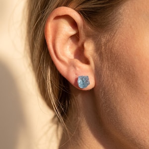 Könnte beinhalten: Ein einzelner Ohrstecker mit einem rohen, ungeschliffenen hellblauen Edelstein in unregelmäßiger, facettierter Form. Der Stein sitzt direkt am Ohrläppchen und zeigt eine durchscheinende, kristalline Textur sowie einen kühlen, hellblauen Farbton.