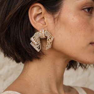Könnte beinhalten: Ein goldfarbener Statement-Ohrring in Schleifenform, besetzt mit einer dichten Anordnung klarer, rechteckiger Baguette-Kristalle. Das Design umfasst ein zentrales Knotendetail mit beidseitig herabhängenden, kristallbesetzten Schlaufen.