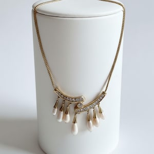 May include: Gold-toned necklace with a delicate chain and a pendant featuring curved bars adorned with clear rhinestones. The pendant also has teardrop-shaped, off-white beads suspended from it. The necklace is displayed on a white cylindrical stand.