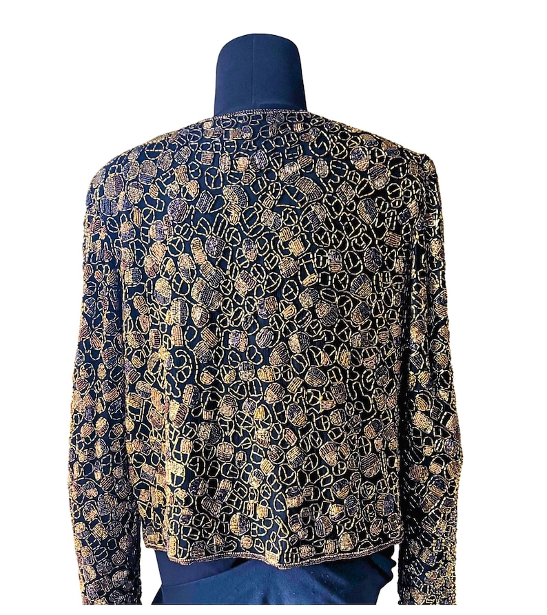 May include: A black jacket with intricate gold and brown beadwork. The jacket is displayed on a mannequin, showcasing the detailed pattern and design. The long sleeves and round neckline are visible.
