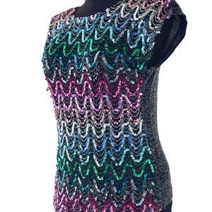 May include: A sleeveless top with a black base and a sequined design in a wavy pattern. The sequins transition through a rainbow of colors, including pink, green, blue, and silver. The top is displayed on a black mannequin.