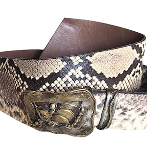 May include: A wide belt with a snakeskin pattern in shades of brown, tan, and cream. The belt features a large, ornate gold-colored buckle with a decorative design. The interior of the belt is a solid brown color.
