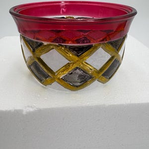 May include: A decorative glass bowl with a ruby red rim and a clear body. The bowl's exterior features a gold and black diamond pattern, adding a touch of elegance. The bowl is designed to hold small items or serve as a decorative piece.