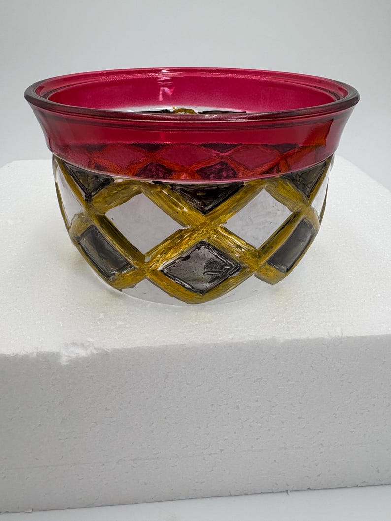 May include: A decorative glass bowl with a ruby red rim and a clear body. The bowl's exterior features a gold and black diamond pattern, adding a touch of elegance. The bowl is designed to hold small items or serve as a decorative piece.