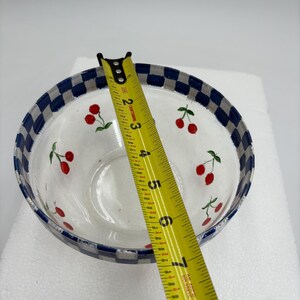 May include: Clear glass bowl with a white interior and a blue and white checkered rim. The bowl is decorated with red cherry illustrations. A yellow tape measure is placed across the bowl, showing a measurement of approximately 20 cm.