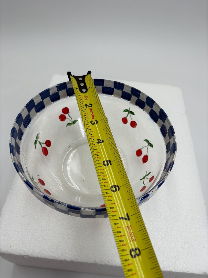May include: Clear glass bowl with a white interior and a blue and white checkered rim. The bowl is decorated with red cherry illustrations. A yellow tape measure is placed across the bowl, showing a measurement of approximately 20 cm.