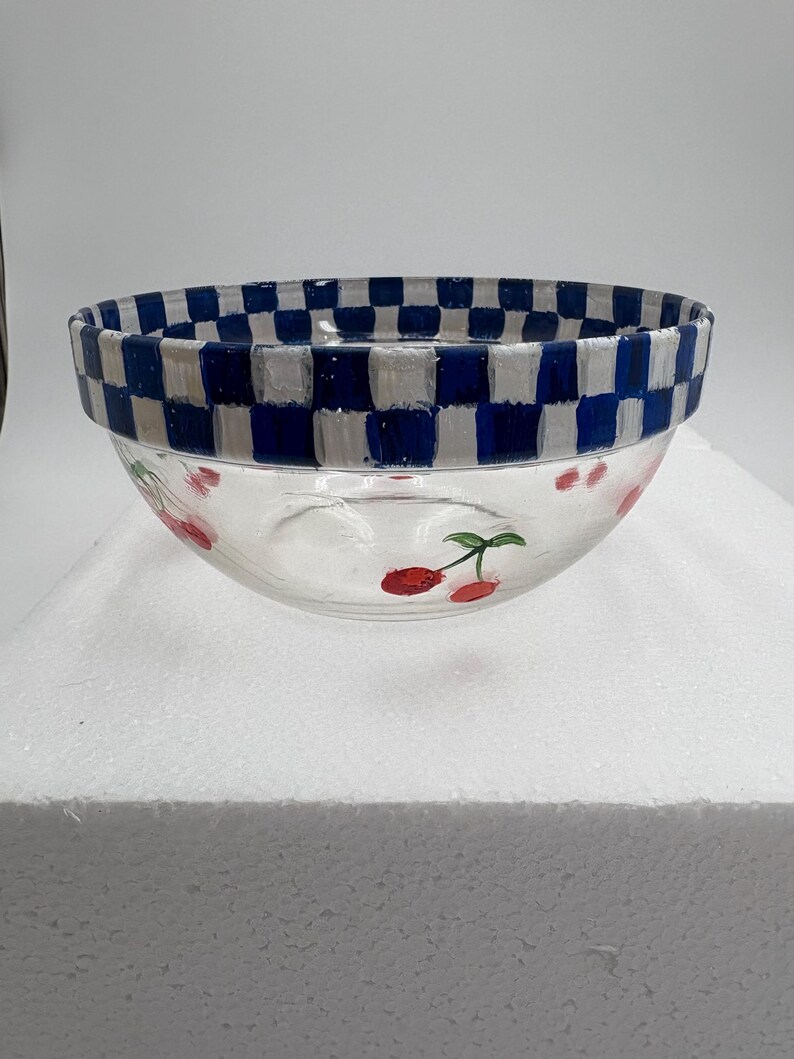 May include: A clear glass bowl with a blue and white chequered rim. The bowl features hand-painted red cherries with green stems. The bowl is a decorative kitchen item.
