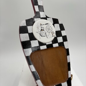 May include: A wooden cutting board with a handle, featuring a black and white checkered pattern. It has a brown rectangular inset and a circular ceramic design with wine-themed illustrations. The board is supported by a gold-colored metal stand.
