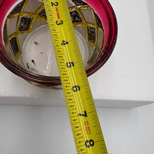 May include: A decorative glass bowl with a red rim and a geometric pattern in black, gold, and gray. The bowl is approximately 4 inches in diameter. A yellow measuring tape is placed across the bowl.