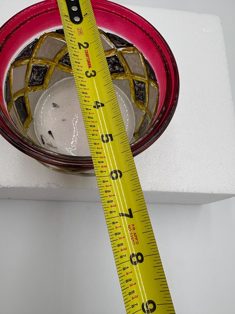May include: A decorative glass bowl with a red rim and a geometric pattern in black, gold, and gray. The bowl is approximately 4 inches in diameter. A yellow measuring tape is placed across the bowl.