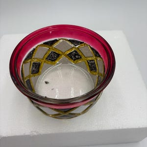 May include: A decorative glass bowl with a vibrant magenta rim. The bowl's exterior features a geometric pattern in gold, black, and gray. The bowl is transparent, allowing a view of its contents. The design is intricate and eye-catching.