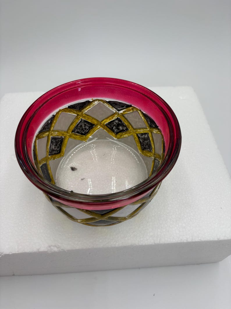 May include: A decorative glass bowl with a vibrant magenta rim. The bowl's exterior features a geometric pattern in gold, black, and gray. The bowl is transparent, allowing a view of its contents. The design is intricate and eye-catching.