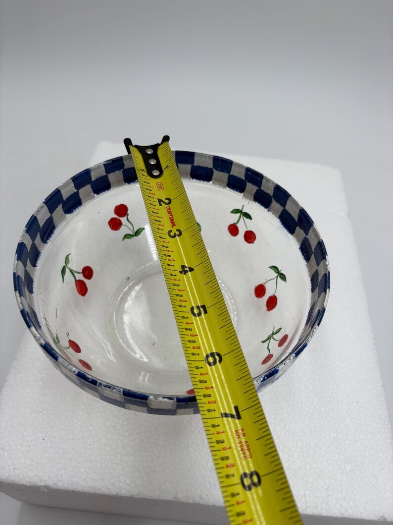 May include: A clear glass bowl with a blue and white checkered rim. The bowl is decorated with red cherries and green leaves. The bowl is approximately 18 cm in diameter.