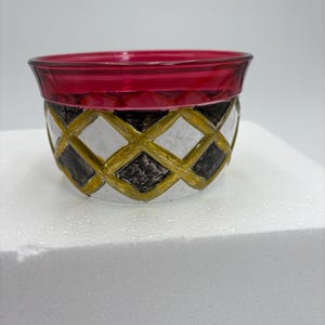 May include: A decorative glass bowl with a red rim and a patterned exterior. The pattern features a diamond design in gold, black, and white. The bowl is sitting on a white surface.