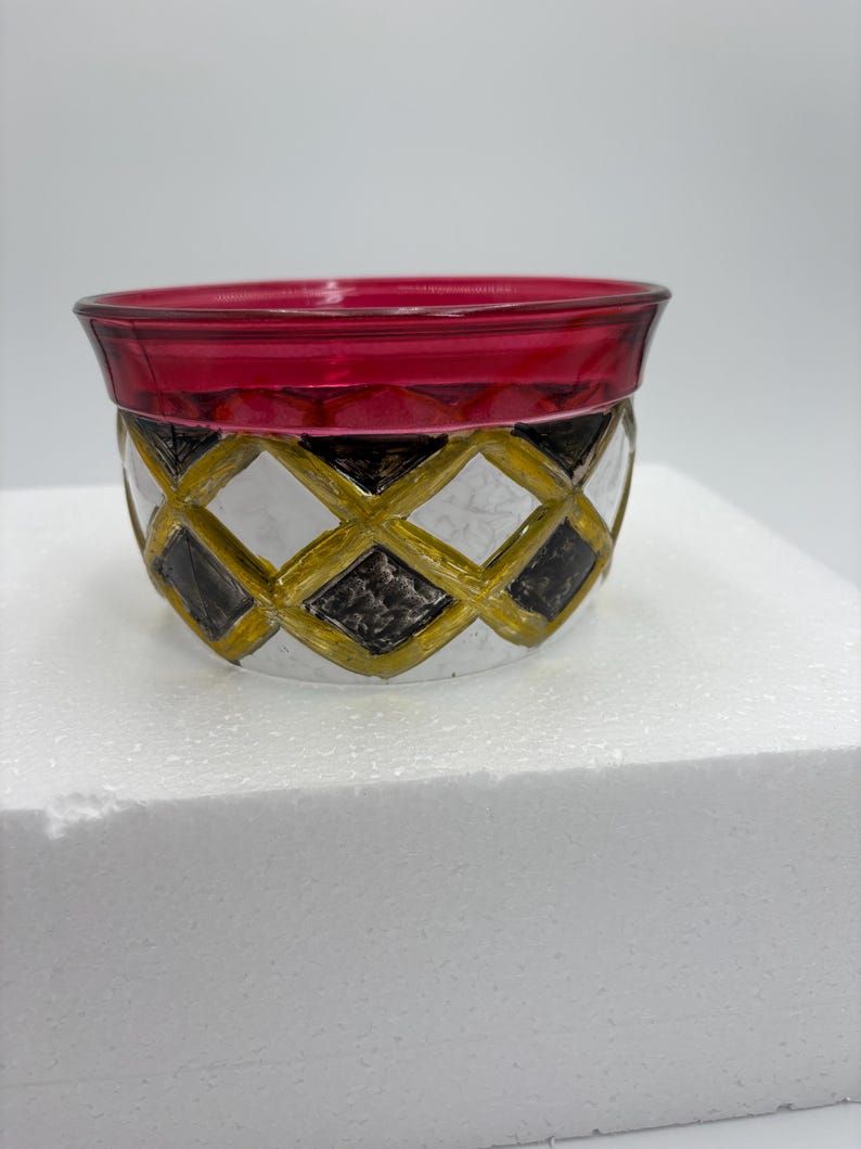 May include: A decorative glass bowl with a red rim and a patterned exterior. The pattern features a diamond design in gold, black, and white. The bowl is sitting on a white surface.