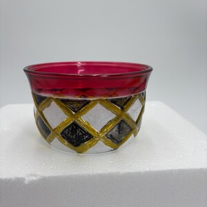 May include: A small, round glass bowl with a ruby red rim and a geometric pattern. The pattern features black, white, and gold diamond shapes. The bowl sits on a white surface.