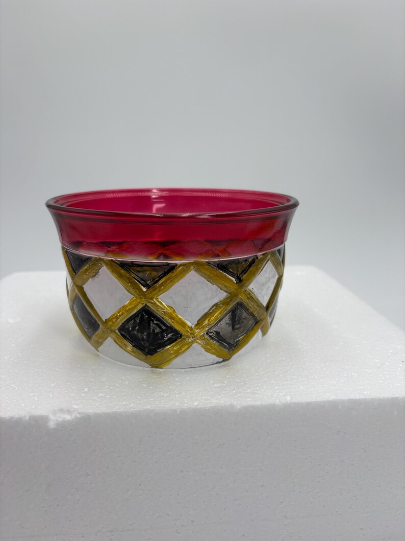 May include: A small, round glass bowl with a ruby red rim and a geometric pattern. The pattern features black, white, and gold diamond shapes. The bowl sits on a white surface.