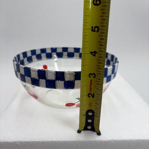 May include: A clear glass bowl with a blue and white chequered rim and red cherry designs. The bowl is approximately 10 cm in height. The bowl is sitting on a white surface.